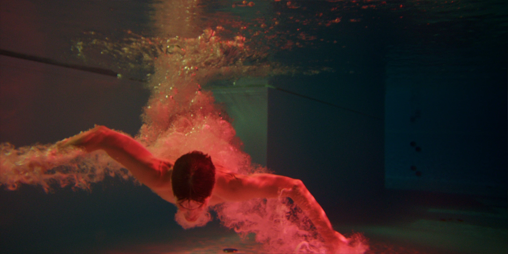 Louis (Benjamin Lillie) dives into a greenish pool like a bird. The air bubbles created illuminate red.