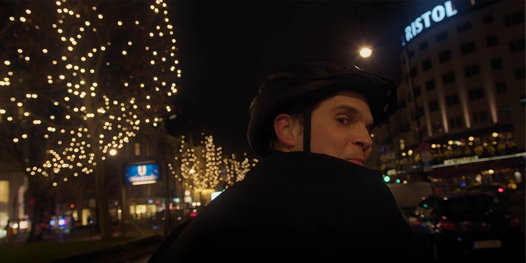 A young man (Benjamin Lillie) rides a tandem bike on Kudamm. He looks towards the camera. The trees on the median strip are illuminated with Christmas lights in the TV commercial »True Romance«.