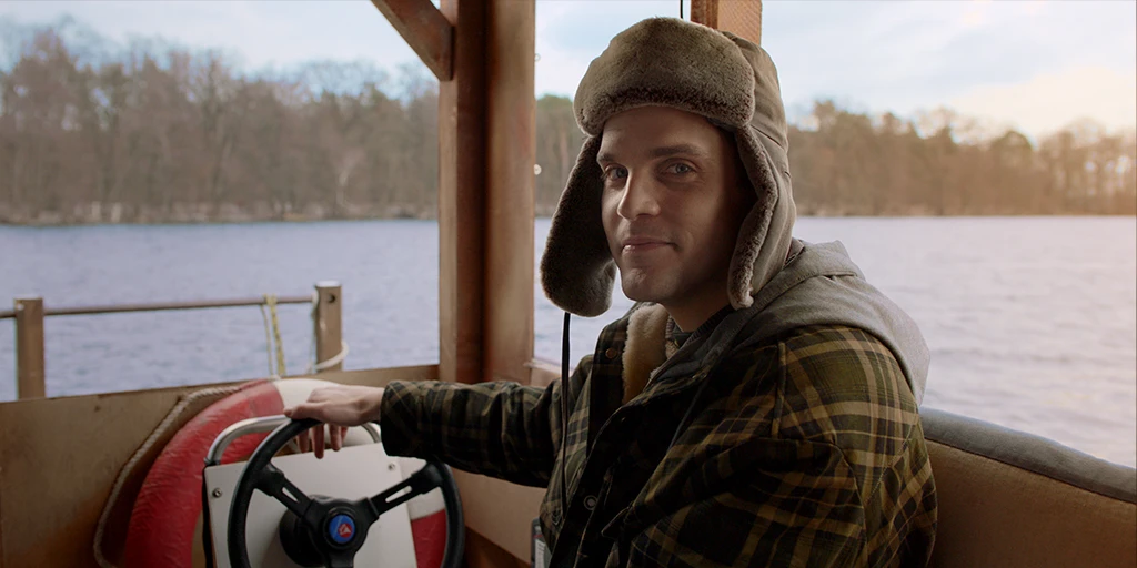 A young man (Benjamin Lillie) steers a wooden raft across an autumnal lake. He wears a warm hat. His expression is expectant in the TV commercial »True Romance«.