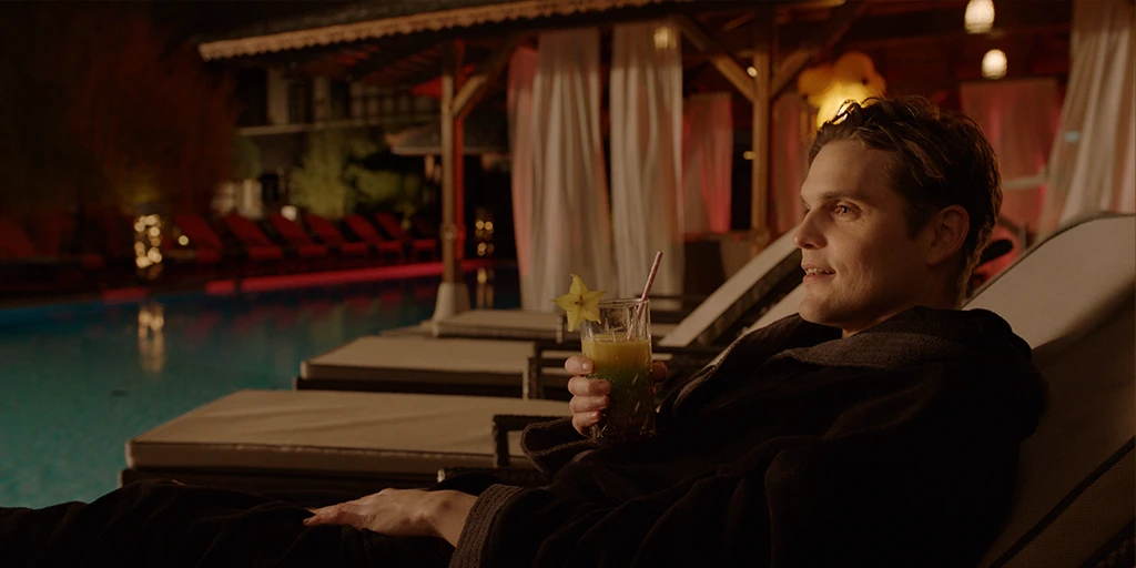 Louis (Benjamin Lillie) sits deeply relaxed on a lounger in a spa, sipping a cocktail. He wears a brown bathrobe and enjoys a cozy atmosphere in the TV commercial »True Romance«.