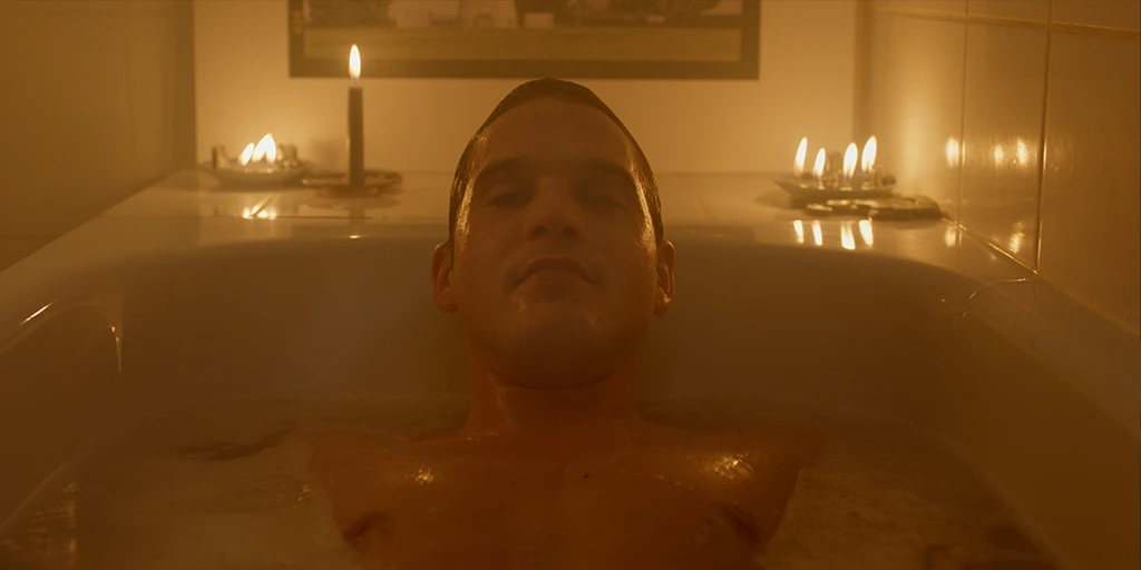 Louis (Benjamin Lillie) lies in the bathtub by candlelight and looks at the camera. Behind him is an art photograph in the TV commercial »True Romance«.