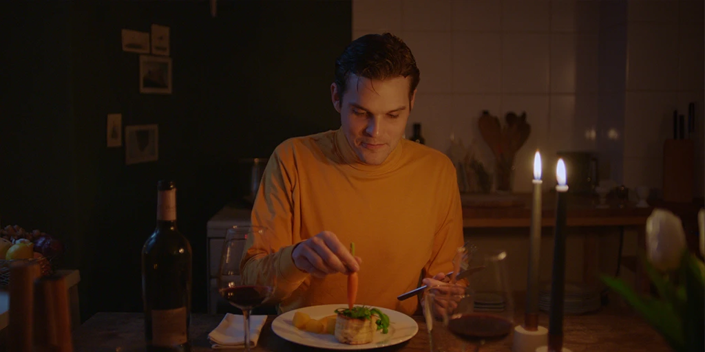 He (Benjamin Lillie) has prepared a stylish dinner for her. He sits in a yellow top in his kitchen. There is wine on the table. He dips a carrot into a mini quiche in the TV commercial »True Romance«.