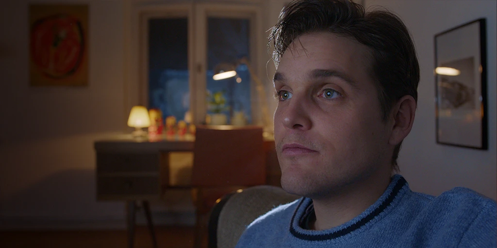 A young man (Benjamin Lillie) watches a movie intently. The light from the screen reflects in his eyes. He wears a blue sweater. In the background, there is a desk with Matryoshka dolls. The view through the window behind reveals a bluish evening atmosphere in the TV commercial »True Romance«.