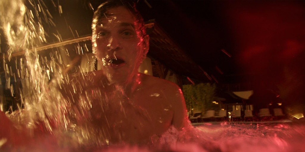 He (Benjamin Lillie) is just emerging from the swimming pool and looks delightedly at the camera. The water is agitated by his emergence. Warm light hits his face from the left. Reddish light illuminates the scene from the right. Behind him is a pavilion in the TV commercial »True Romance«.