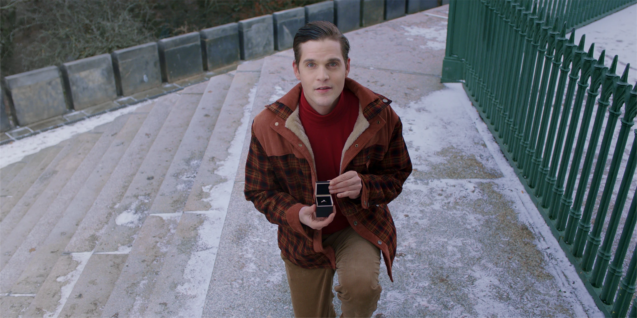 A young man (Benjamin Lillie) kneels and opens a ring box. He is dressed in reddish-brown. Next to him is a green fence. He looks expectantly at the camera, waiting for her to say yes in the TV commercial »True Romance«.