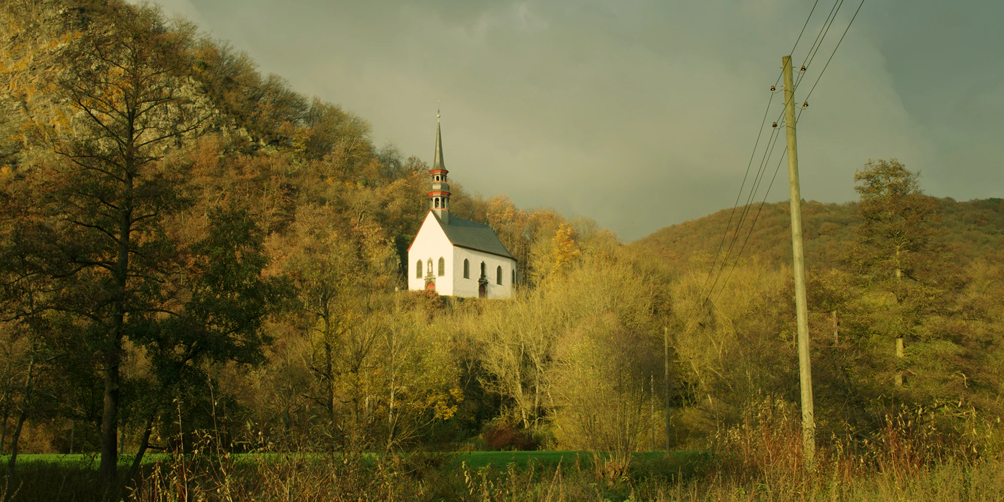 We look up from the valley at a small chapel standing on a plateau of a mountain. Sunlight illuminates the white front in twilight. To the left above the chapel, a steep rock face can be seen. A power line leads towards the chapel.