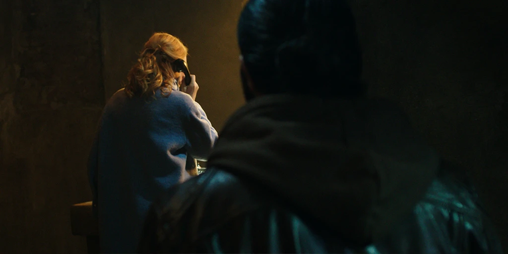 Katja stands at an altar, making a phone call. She wears a blue coat, and warm light hits her side. A man in a brown leather jacket approaches her slowly.