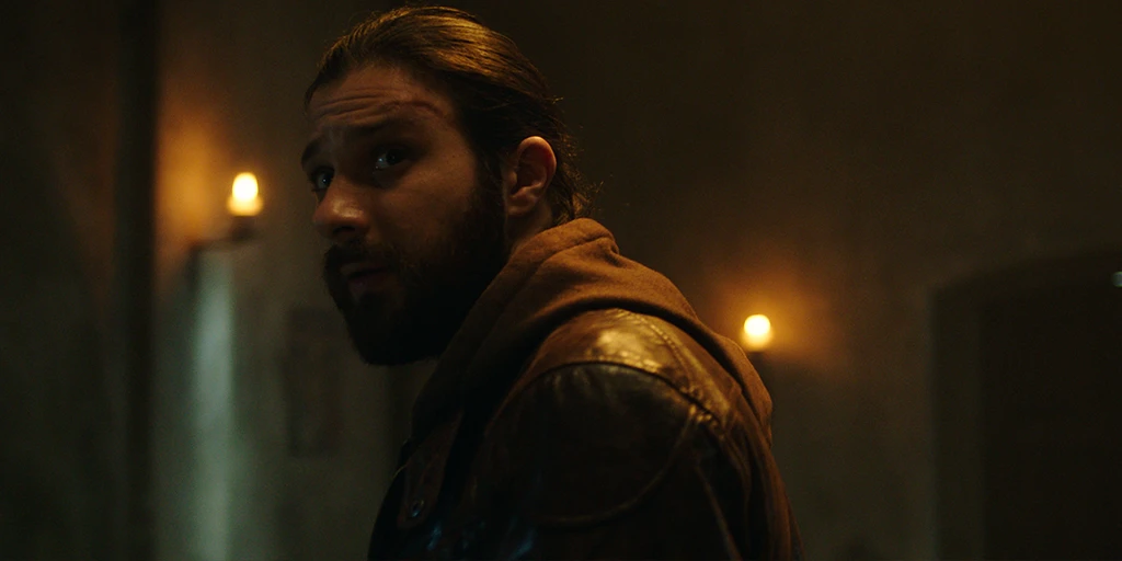 A middle-aged man looks towards the altar in a small chapel. He wears a brown leather jacket. The chapel behind him is illuminated by candles. He looks forward expectantly.