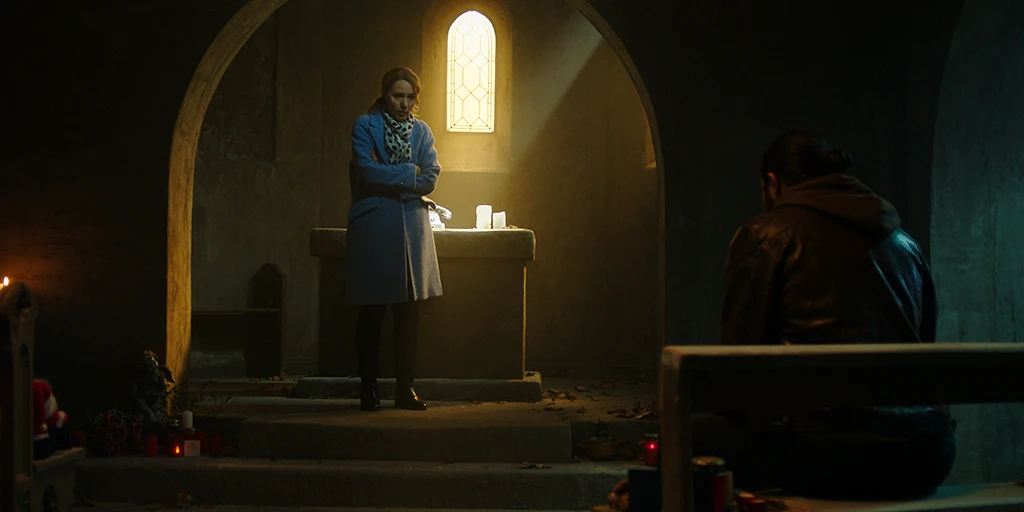 A middle-aged woman stands at an altar. Warm sunlight shines on the altar and the phone placed on it. With folded arms, she talks to a middle-aged man who sits defiantly on one of the chapel benches.