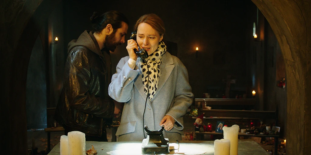 A middle-aged woman stands in front of an altar, making a phone call with the phone placed on it. Visibly emotional, she speaks into the receiver. A man in a brown leather jacket leans in to listen. In the background of the chapel, many memorial candles are set up.