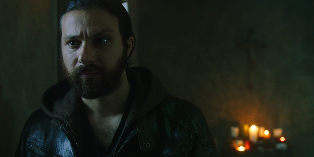 A middle-aged man with black hair and a brown leather jacket looks disappointedly out of the frame. Behind him is a cross and a table with lit memorial candles. Daylight from a chapel window shines on him from the side.