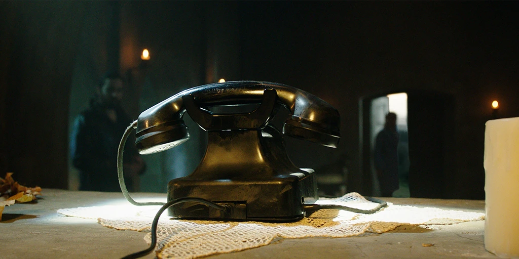 A black rotary phone stands in the middle of the altar. In the background, a man looks at the phone in amazement. A middle-aged woman stands at the open chapel door, also looking towards the rotary phone.