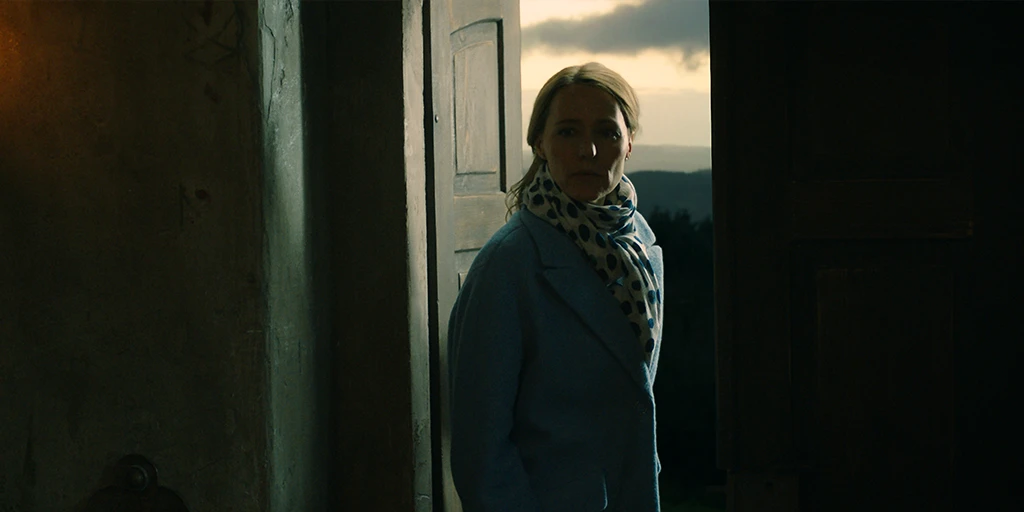 A middle-aged woman stands at an open chapel door. Her gaze is turned into the chapel. The door is open, and a vast mountain landscape can be seen in the background.