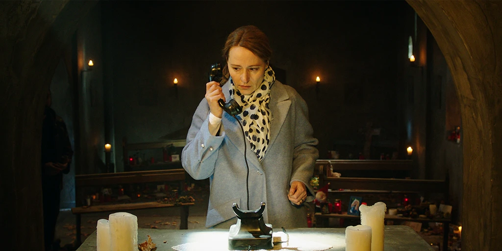 A woman with blonde hair stands in front of an altar and holds the receiver of a black rotary phone to her ear. In the background, many memorial candles are placed on the church benches. Behind the chapel arch, the figure of a man can be seen.