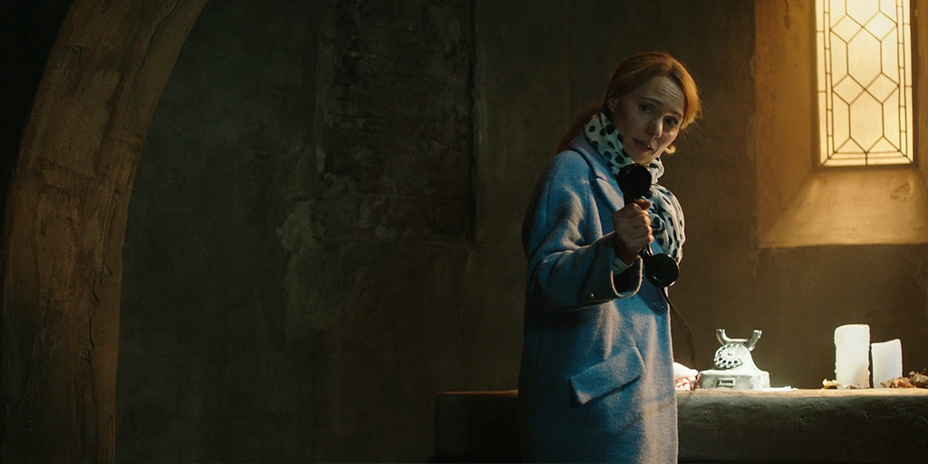 A woman with blonde hair holds the receiver of a rotary phone in her hand, which stands on the altar behind her. She is in the altar room of a small chapel. On the altar, besides the phone, are also two candles and some foliage.