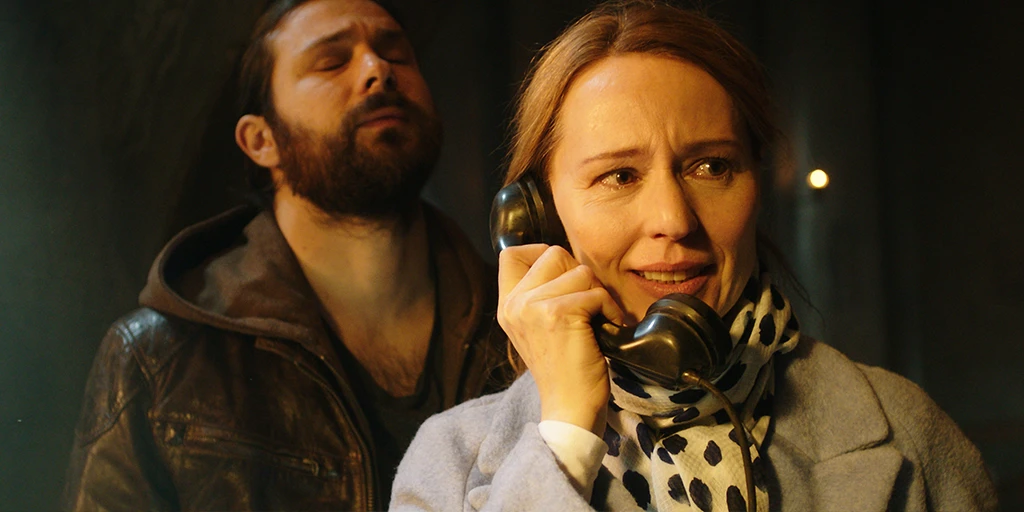 In the foreground on the right stands one of the film's protagonists, Katja. She holds a black receiver to her ear and becomes emotional. To the left behind her stands a man in a brown leather jacket. He tilts his head slightly upward, closes his eyes, and furrows his brow. Both are in the altar room of a small chapel.