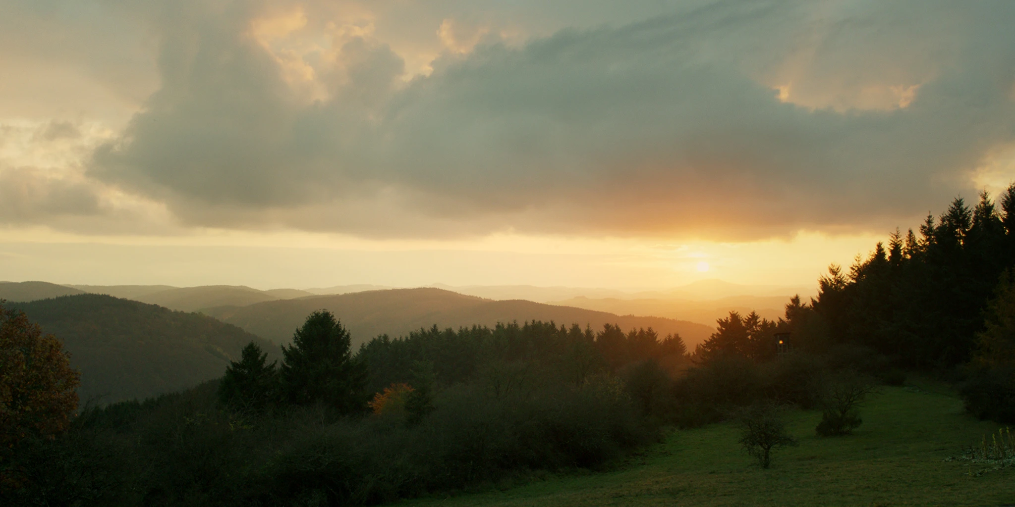 A vast mountain landscape can be seen. The setting sun colors the sky and the mountains yellow. The sun is about to disappear behind one of the mountains. At the edge of the forest, a hunting stand can be seen.